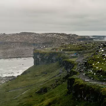 People in the far distance on a path atop a grass-covered rocky hill with water and other rocky hills beyond on a misty, overcast day.