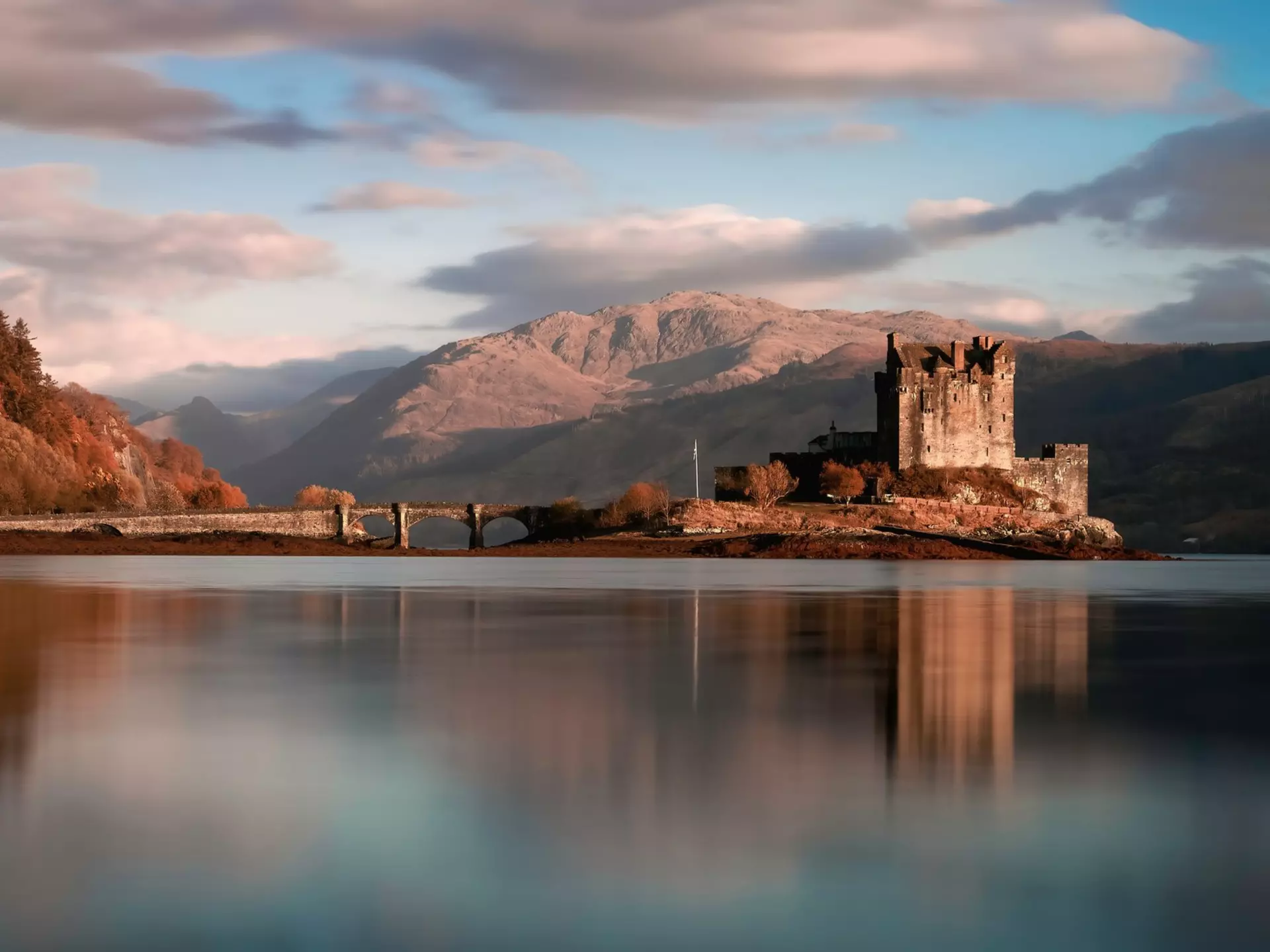 An ancient ruined castle and an arched stone bridge in a lake surrounded by mountains.