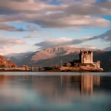An ancient ruined castle and an arched stone bridge in a lake surrounded by mountains.