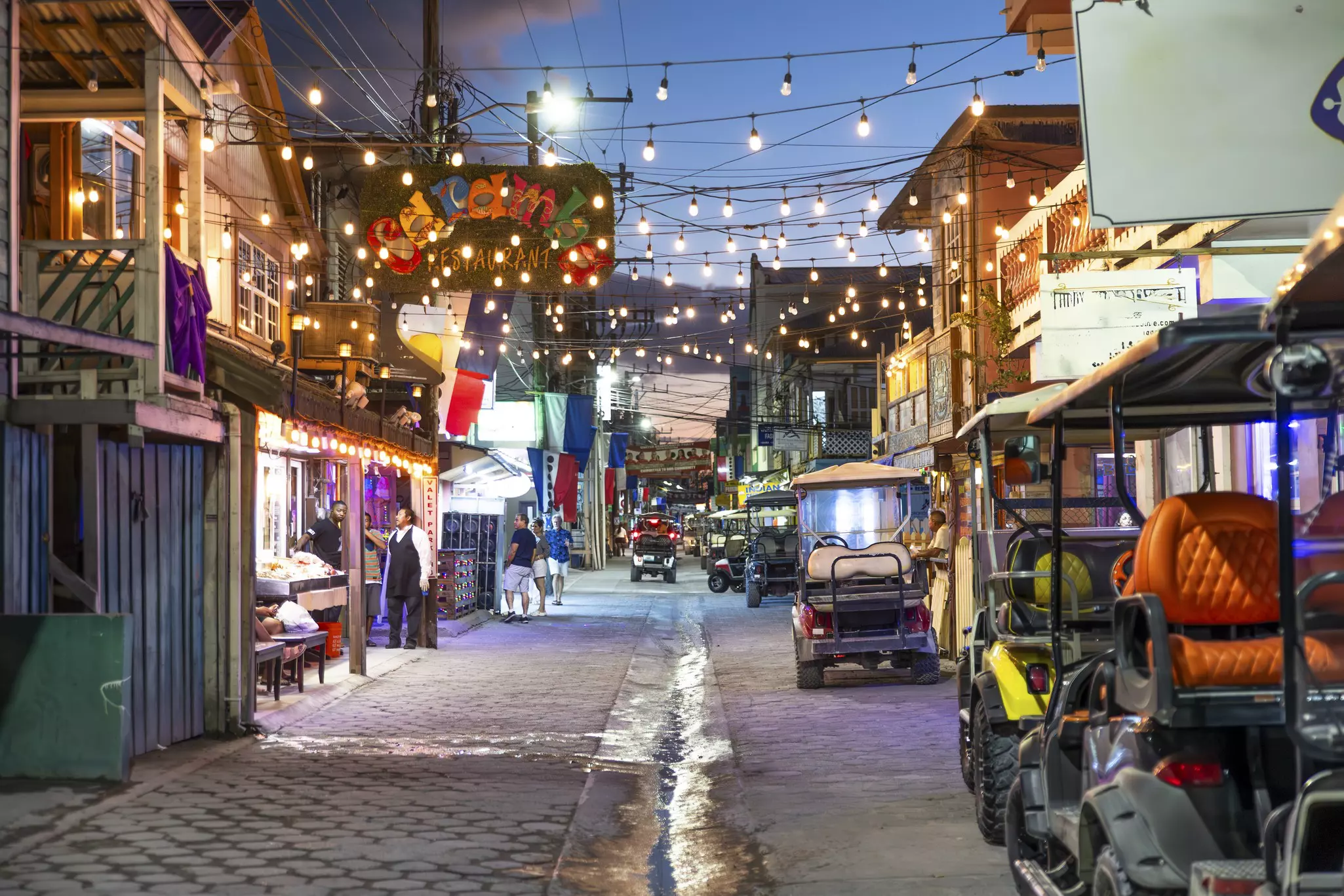 The street of a town is seen in the evening. Lights are strewn over the street, which is filled with golf carts.