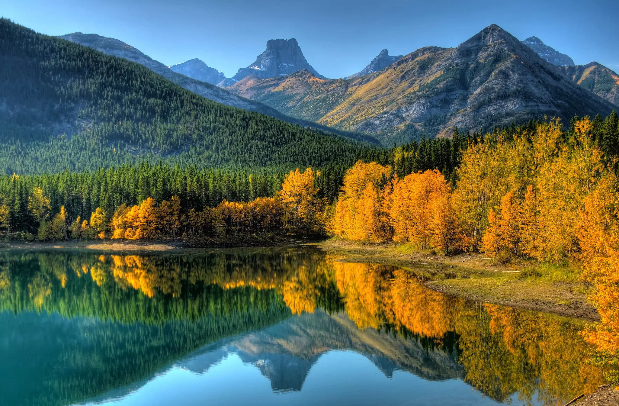 Golden trees by a lake are reflected in the water's surface along with the evergreen-covered mountains in the background.