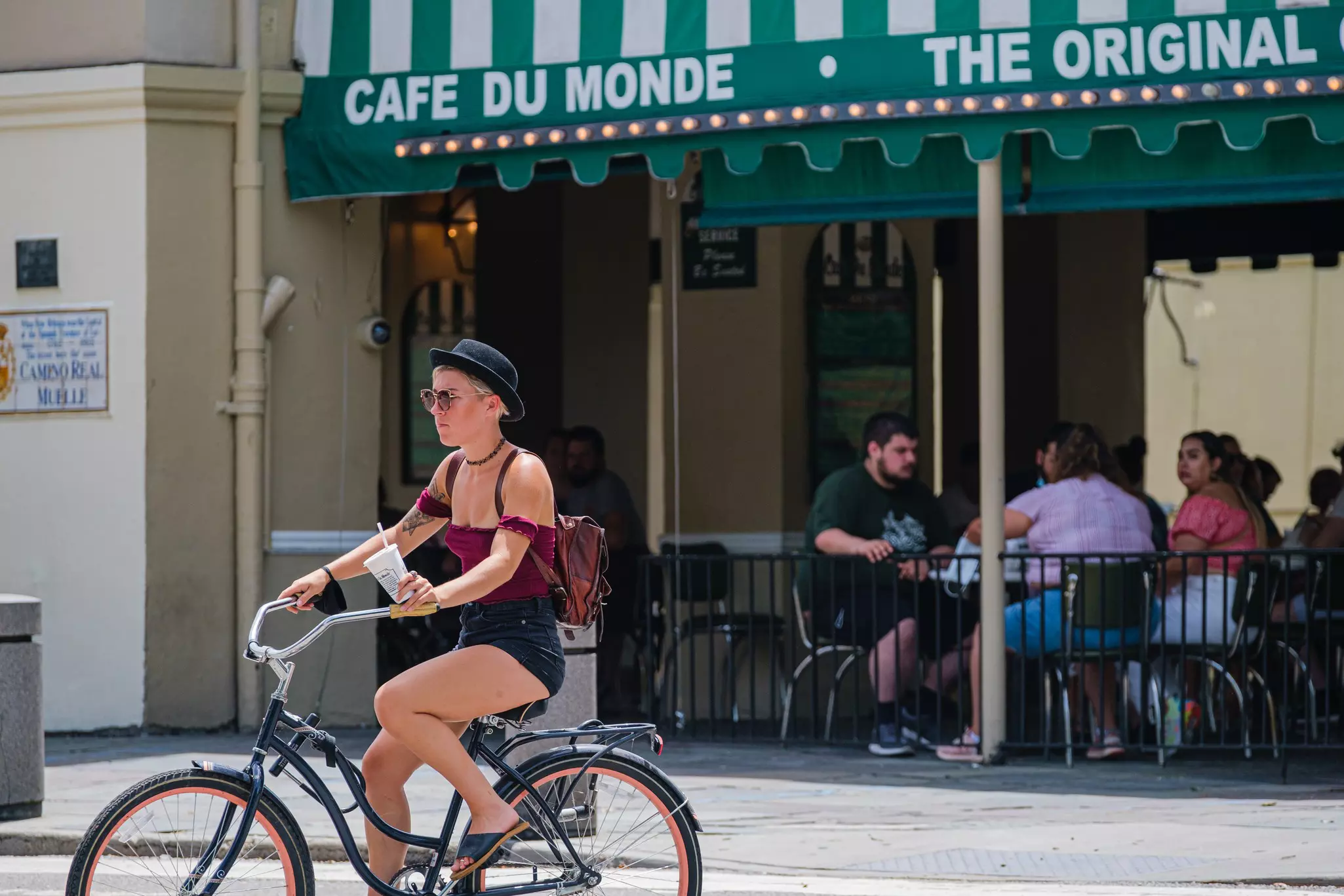 A female cyclist in a black hat pedals past a cafe with outdoor tables under a green awning.