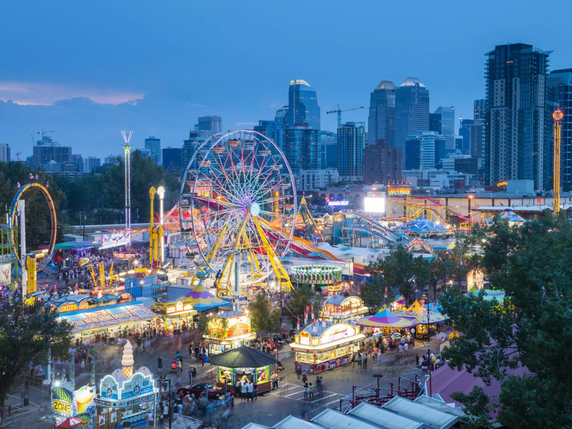 A fun fair with a Ferris wheel lit up at night at a busy event in a city.