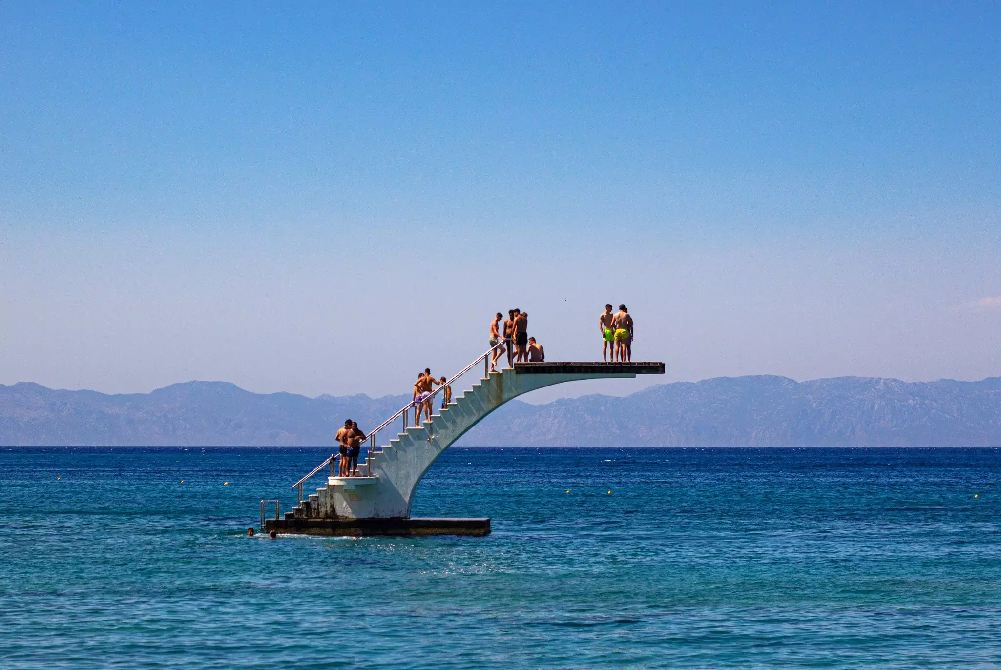 A group of people line up on a diving board that's floating out to sea