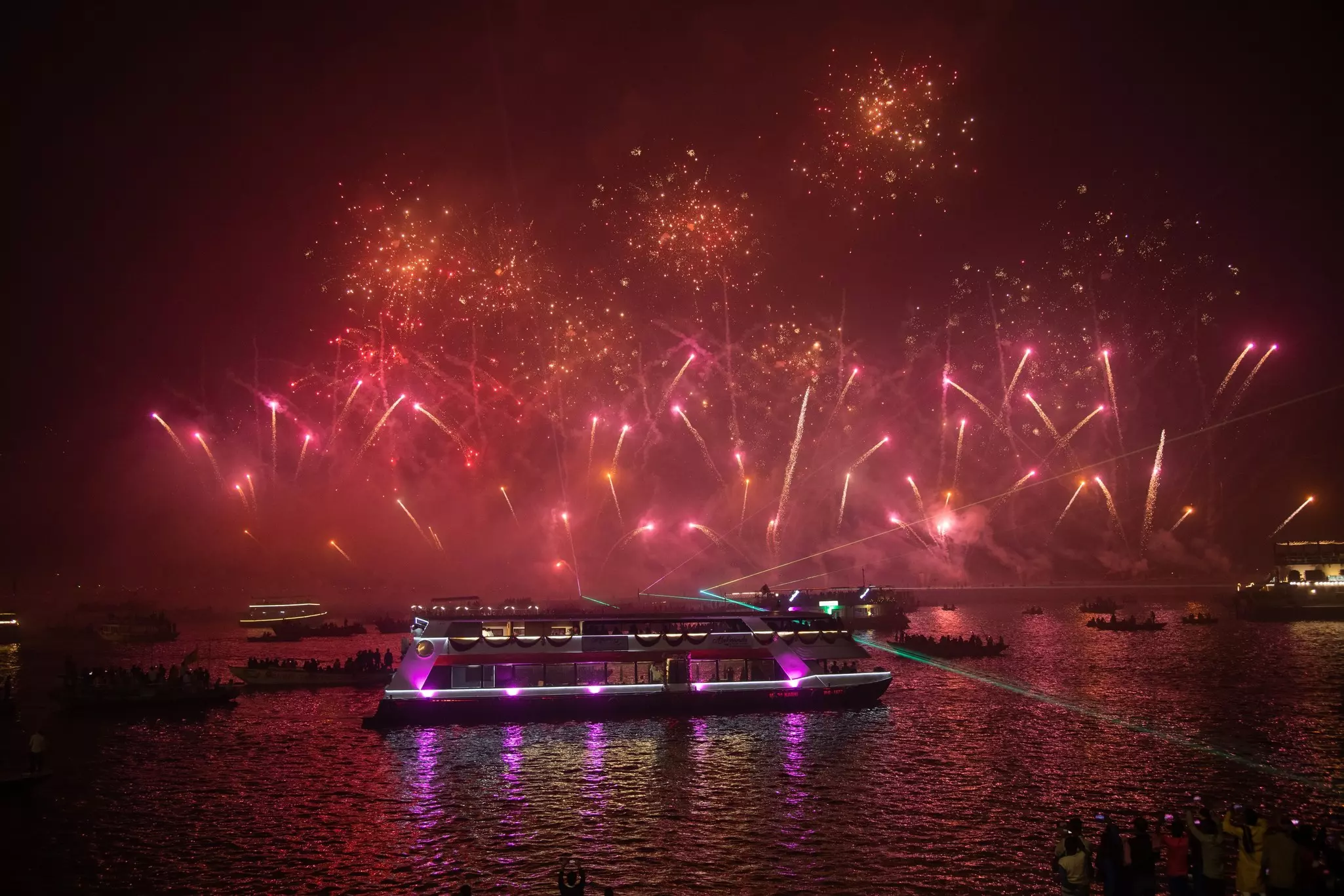 Red-tinted fireworks explode over boats in a river.