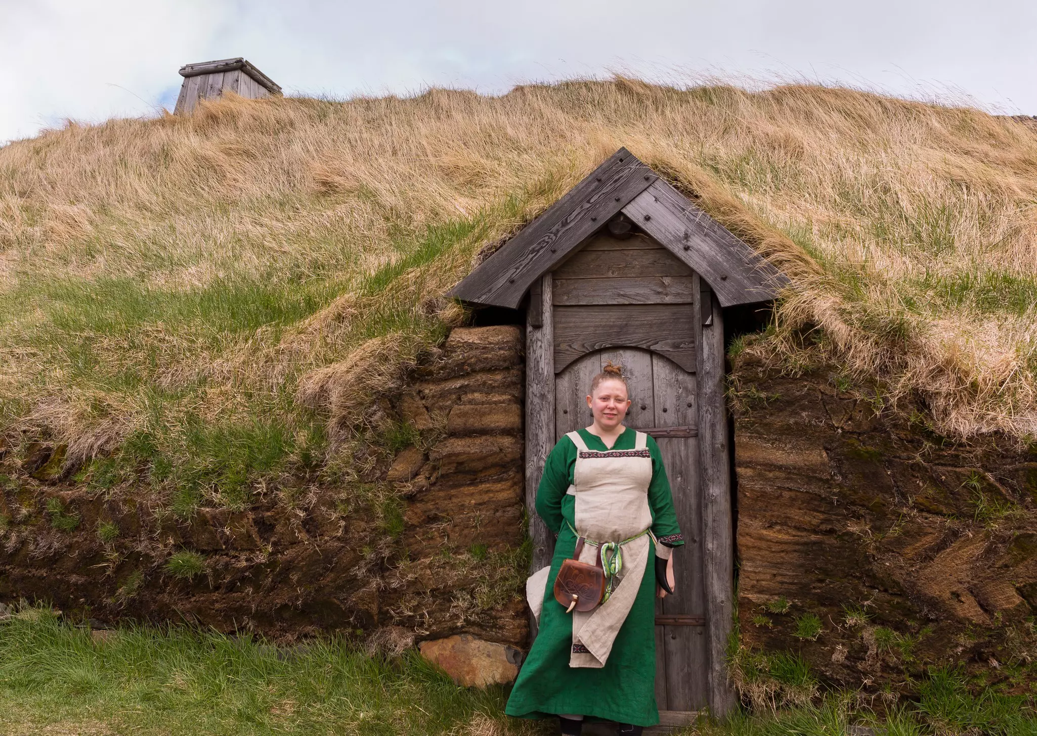 A woman in a green dress and period costume stands in front of a wooden door to a structure covered whose roof is earth and grass.