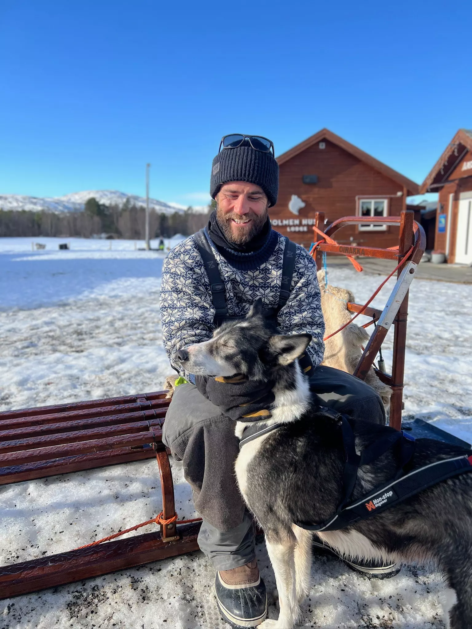 A man in a blue patterned sweater holds a dog outside by a snow-covered field.