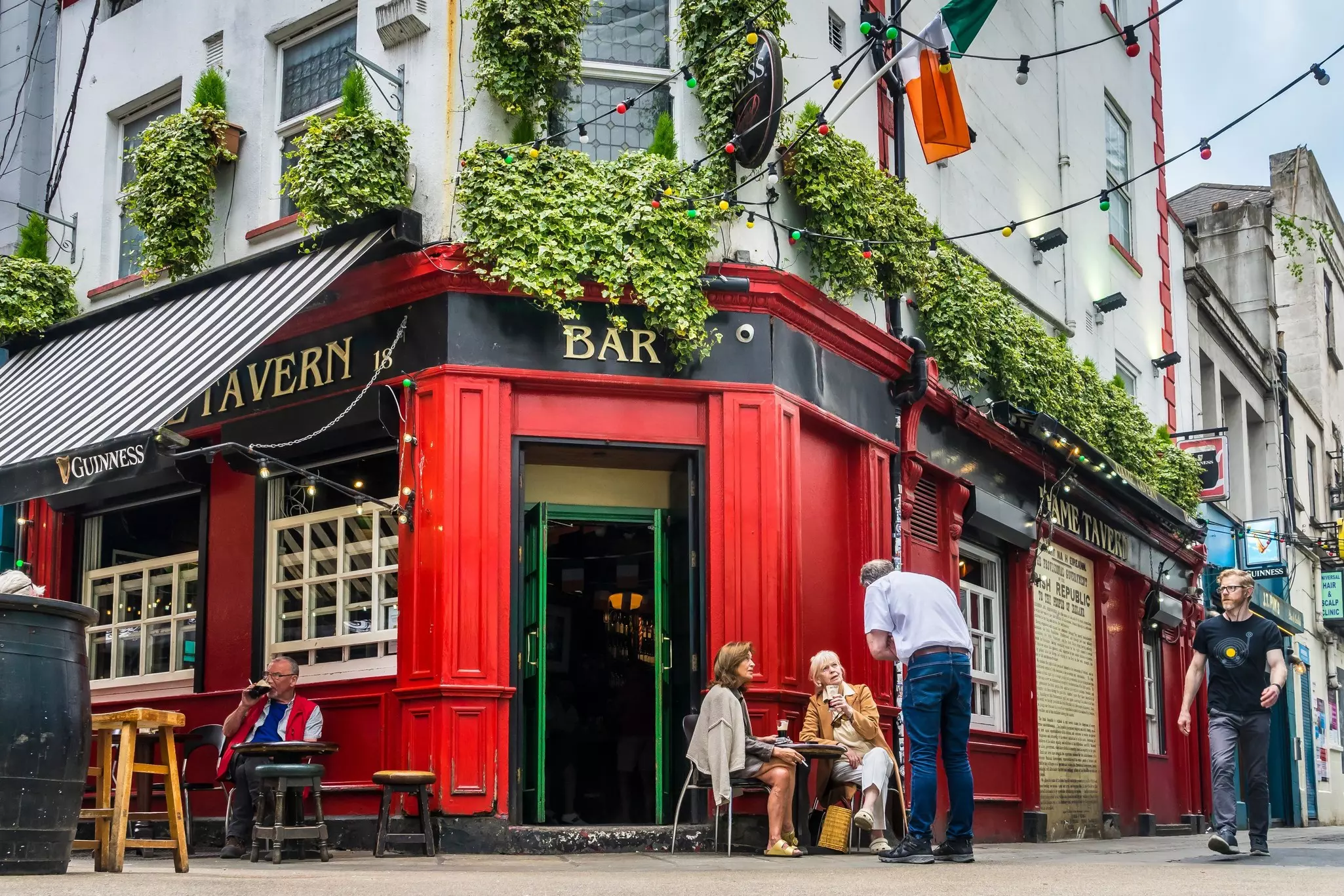 Two people sit at an outdoor table by the doorway to a red pub.