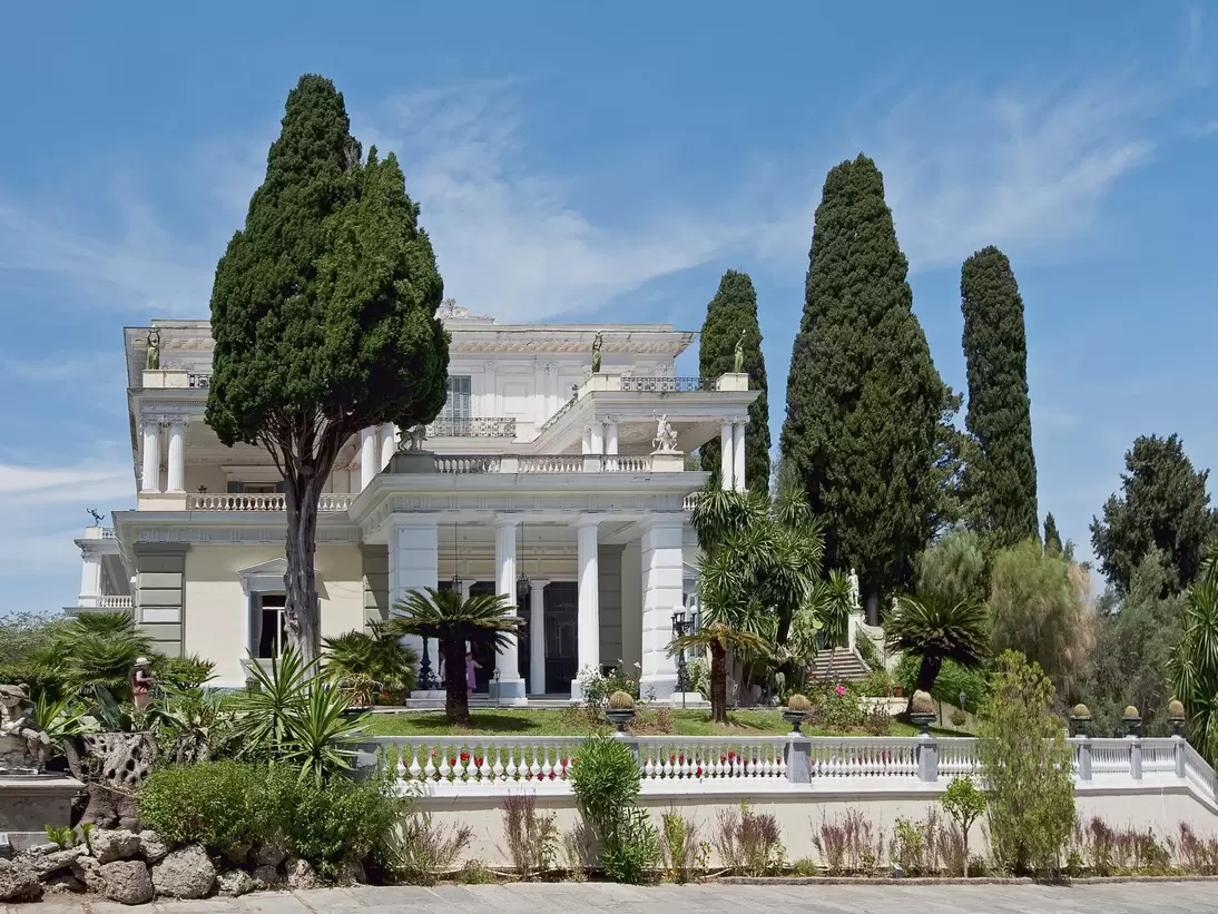 A white neoclassical villa with columned loggias and statues along its roofline, framed by tall cypresses above a balustraded garden terrace.