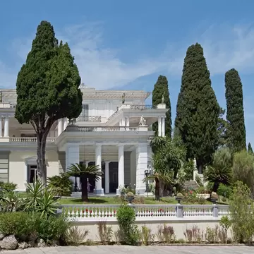 A white neoclassical villa with columned loggias and statues along its roofline, framed by tall cypresses above a balustraded garden terrace.