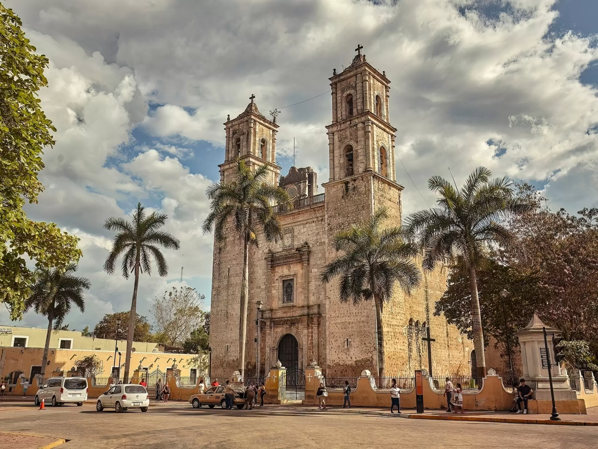 A historic stone church with two towers sits on a city square. A row of palm trees is in front of the church.
