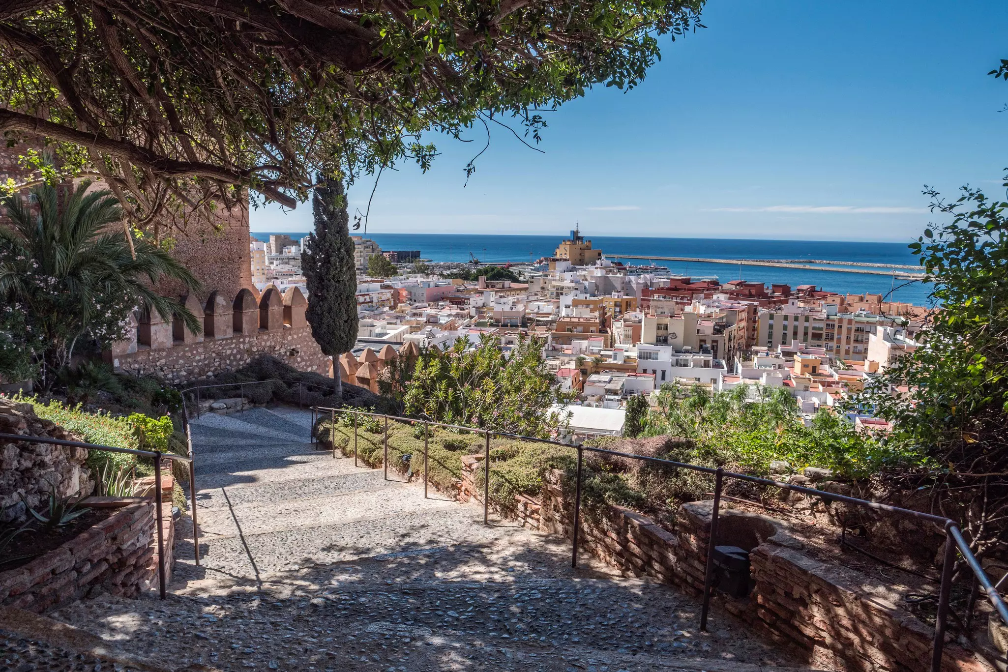 A path weaves downhill in a garden beside a fortress with views stretching over the town and to the ocean