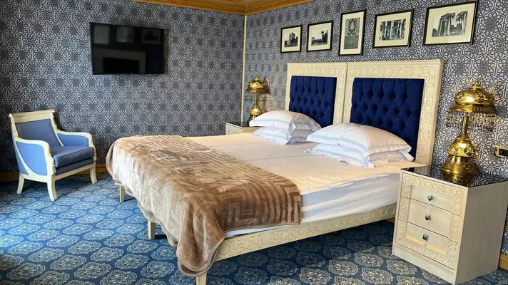Blue pattered wallpaper covers the walls of a room equipped with a large bed with a blue cushioned headboard. Two gold plated lamps are next to it, and the carpet is also a blue pattern