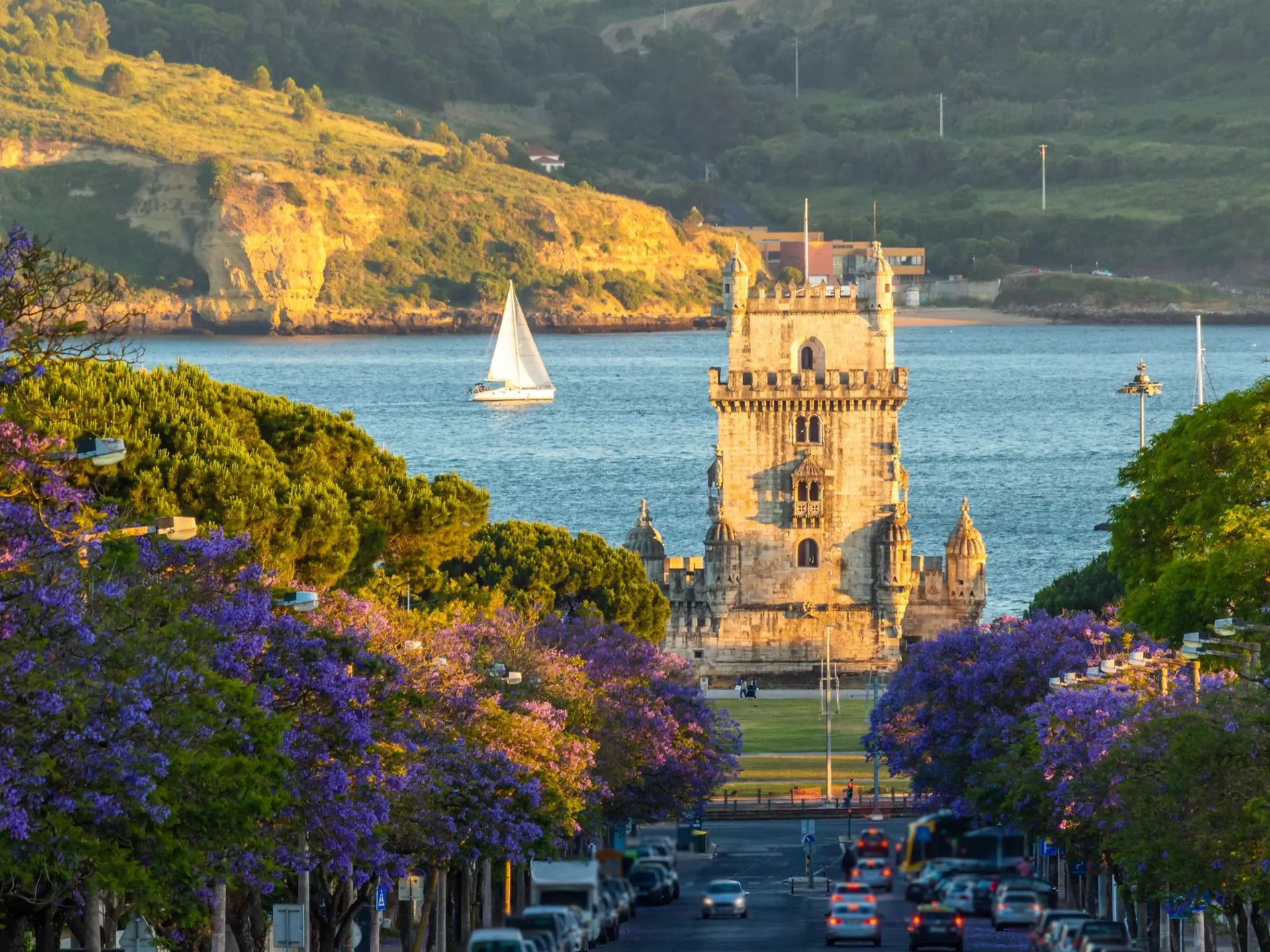 Cars traveling on a street leading to a tower on a shore. Trees with blooming purple flowers line the street. A sailboat is visible on the water.