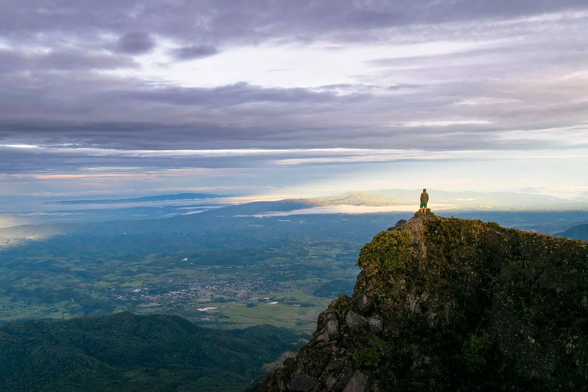 A solo figure stands at a high-altitude rocky outcrop looking over an expanse of land below.