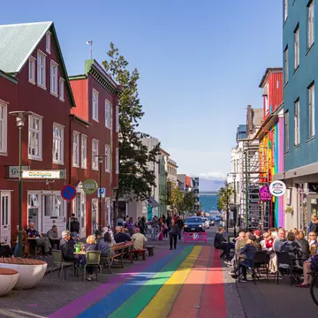 People sit at outdoor cafes on a street with rainbow stripes painted down the middle.