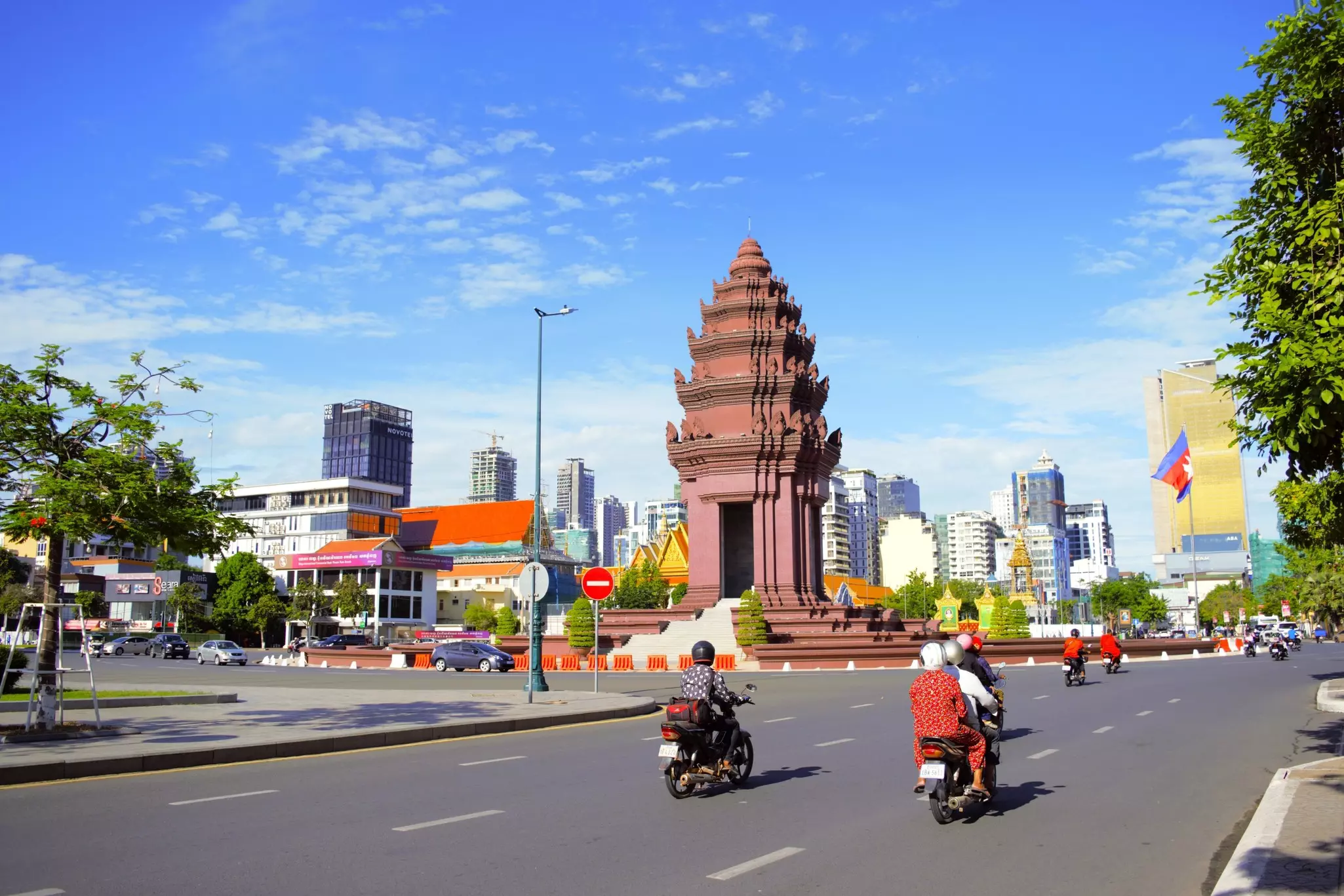 Motorbikes and cars near a traffic circle that has a tall monument in the middle