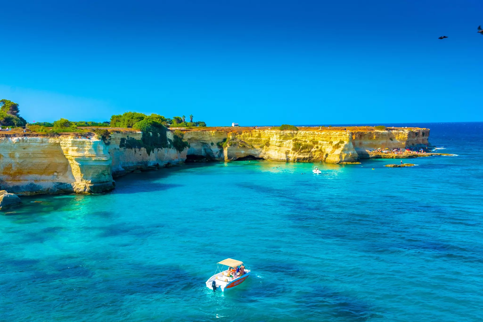 A boat sails by rocky cliffs in Italy; the bright blue water is the same color as the clear sky.