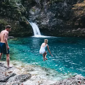 A woman leaps into a clear turquoise plunge pool fed by a waterfall in a rocky gorge as a man watches from the rocks