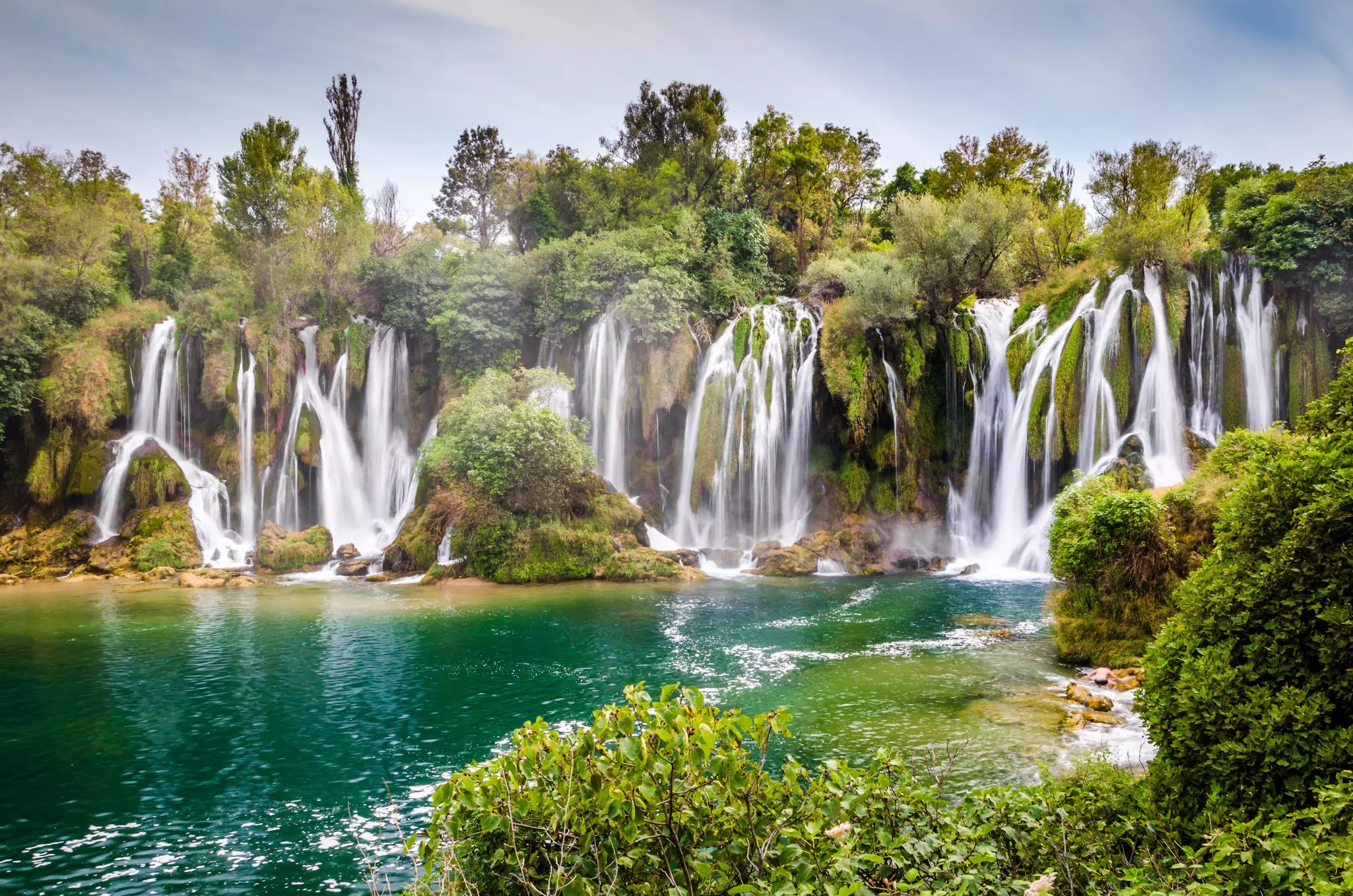 A series of waterfalls plunge down into a green pool in lush woodland.