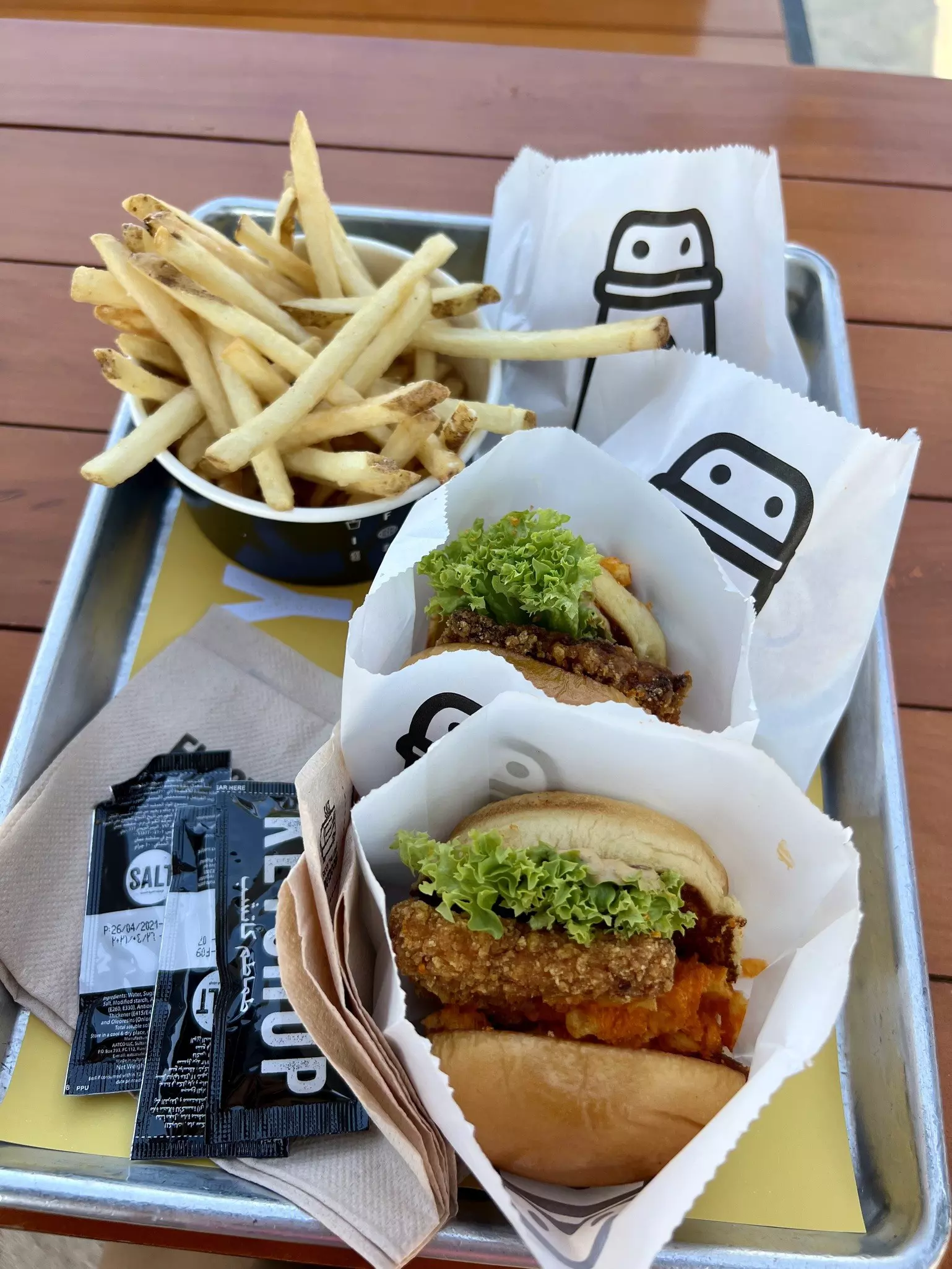 A silver tray with two chicken burgers wrapped in paper alongside a bowl of fries