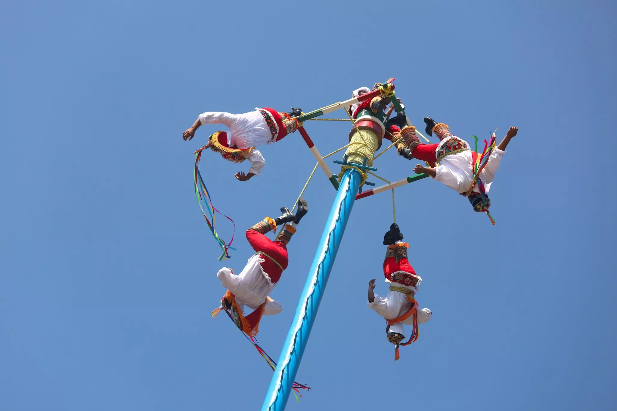 Four men costumed in white shirts and red pants hang by one leg each tied to a high pole on a cloudless day.