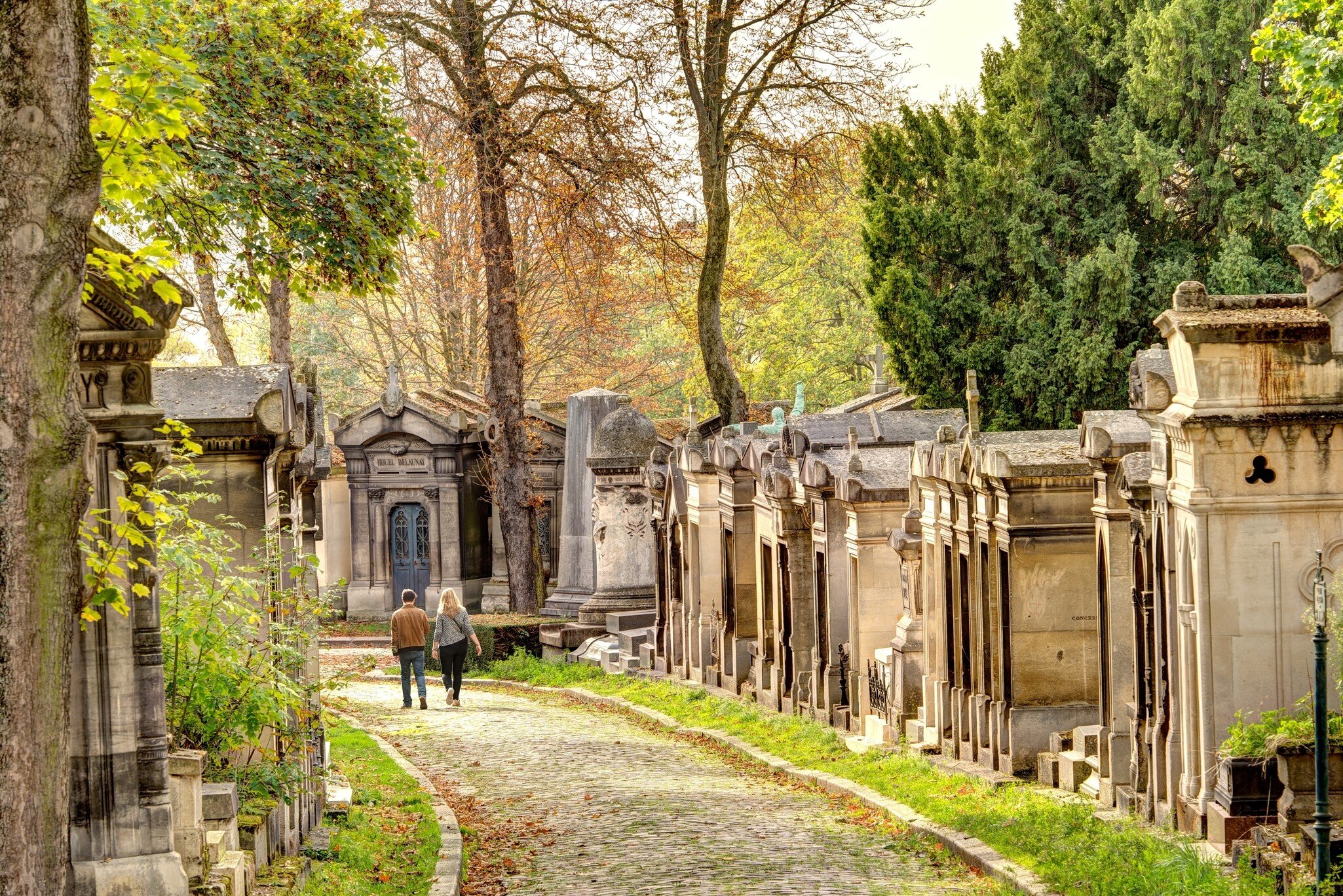 Two people walking on a cobblestone path in a cemetery with mausoleums on either side on an autumnal day.