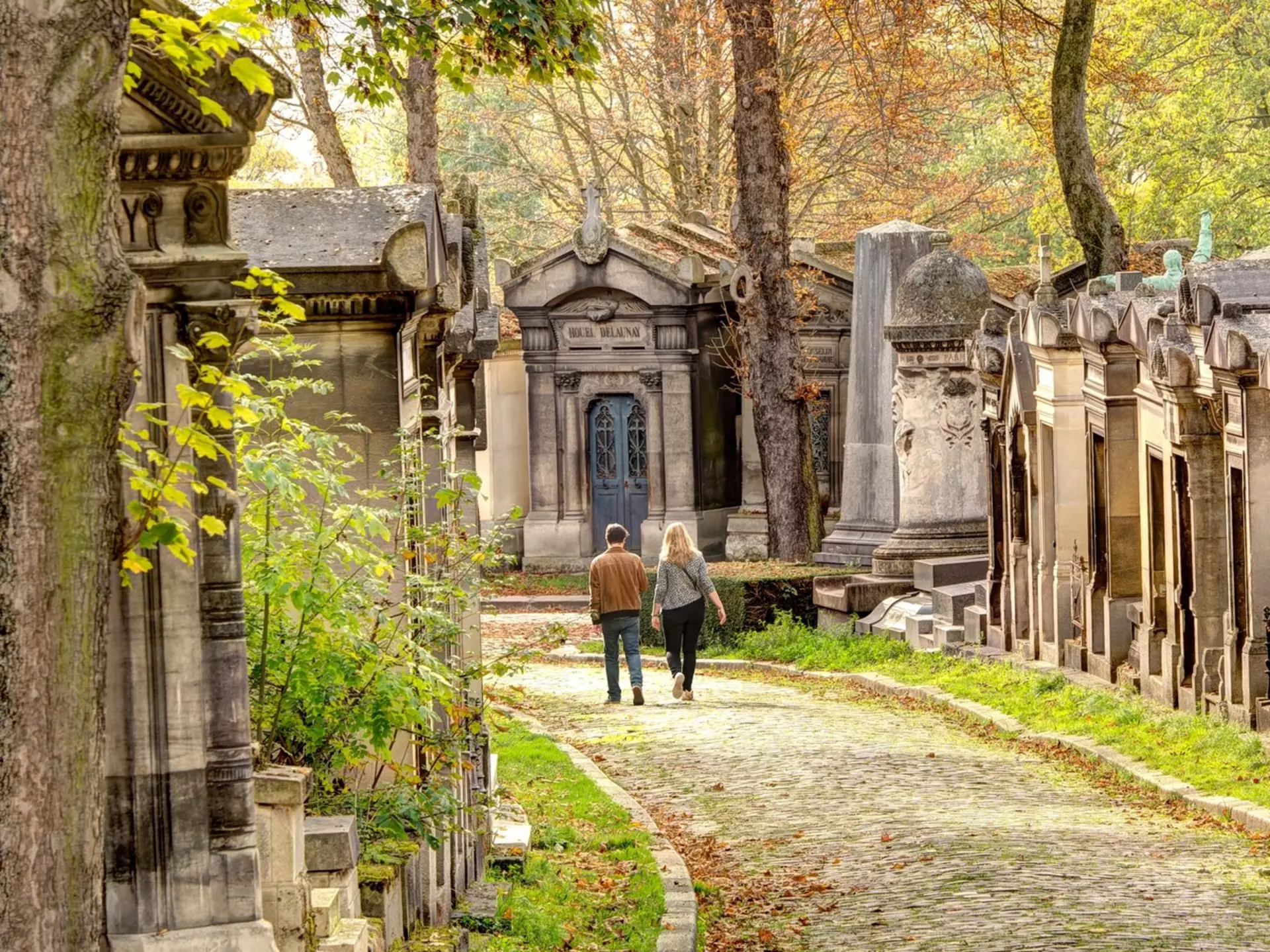 Two people walk down a path lined with mausoleums in autumn, Père Lachaise cemetery, Paris