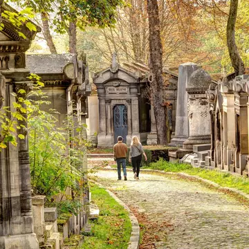Two people walk down a path lined with mausoleums in autumn, Père Lachaise cemetery, Paris