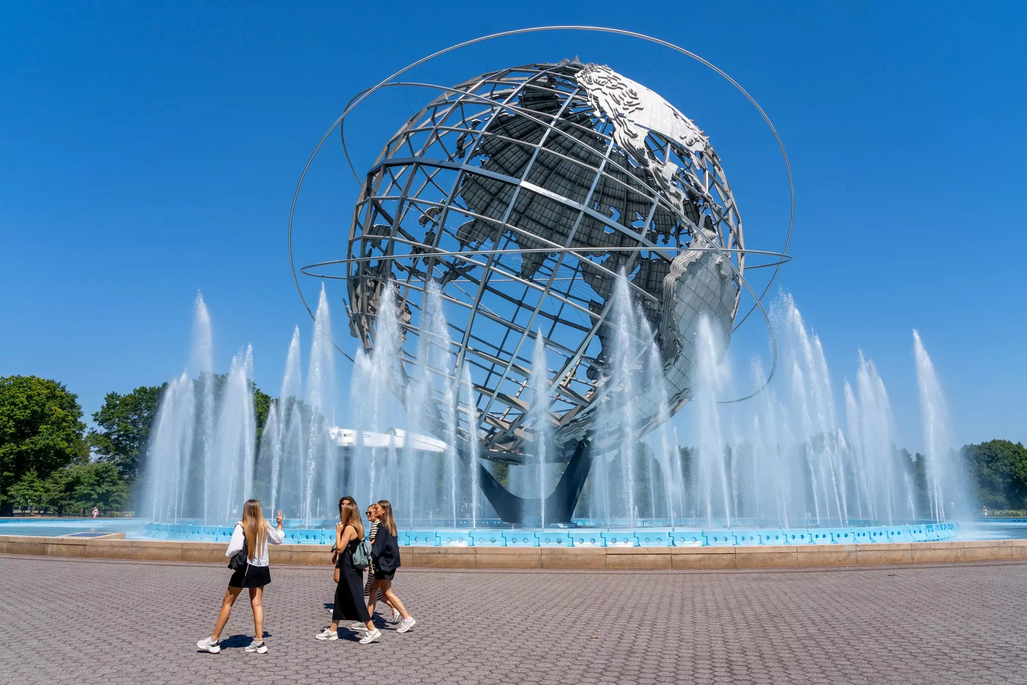 A group of teenagers walks by a giant sculpture in the form of a globe, surrounded by jets of water, in a city park.