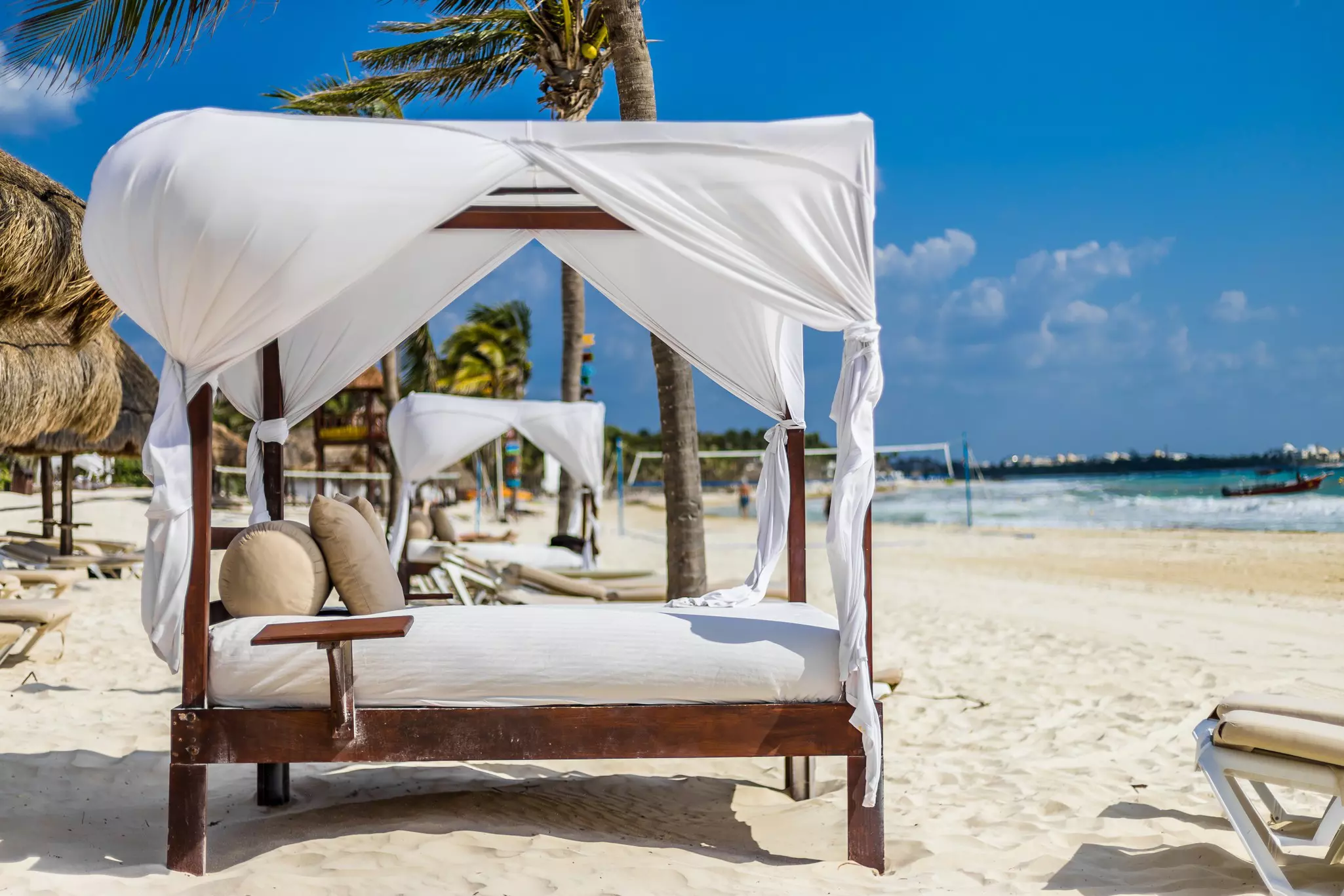 Beds on dark wood frames with white cushions and white curtain canopies are lined up on a white sand beach.