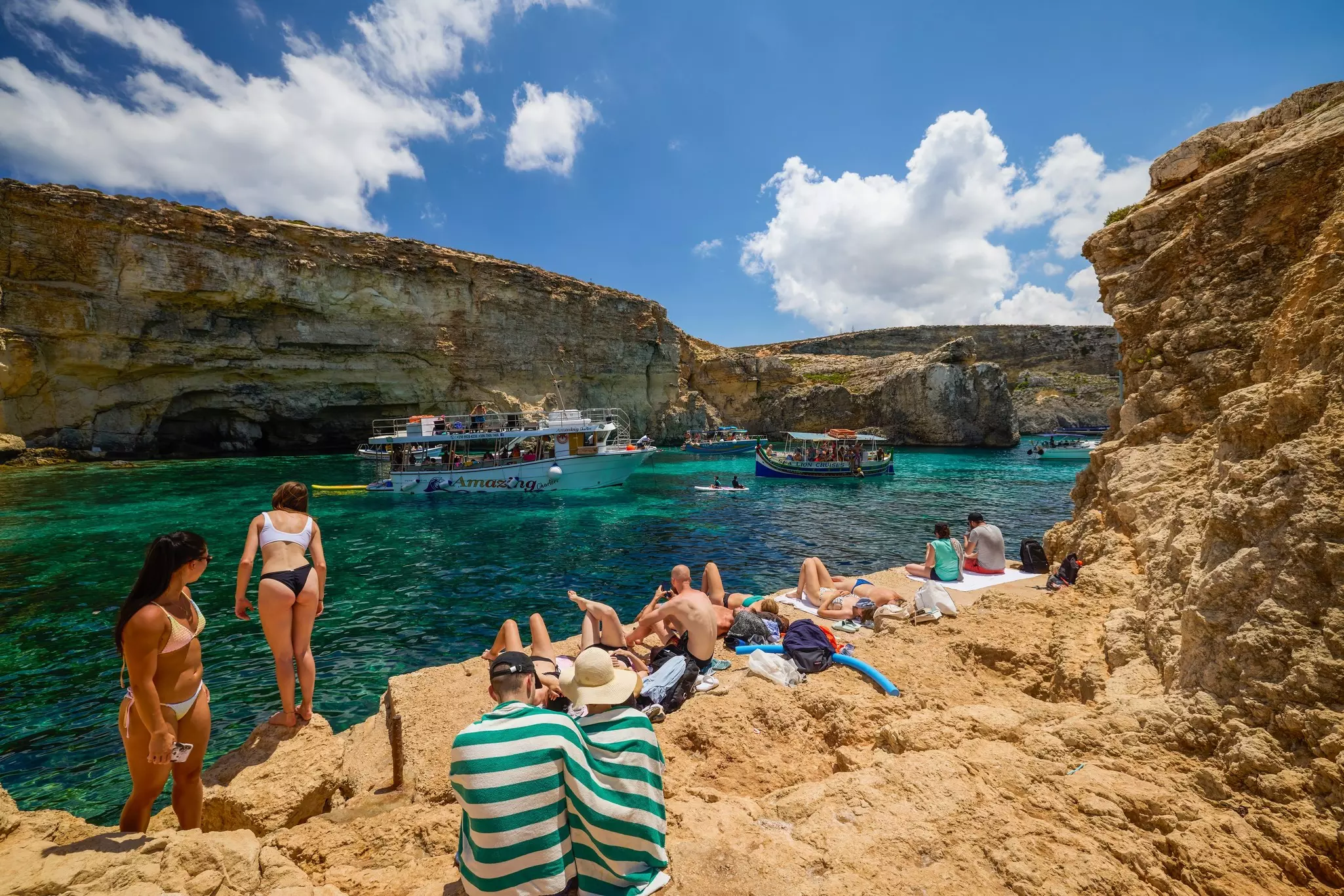People sunbathe on rocks overlooking a lagoon as people in pleasure boats pass by