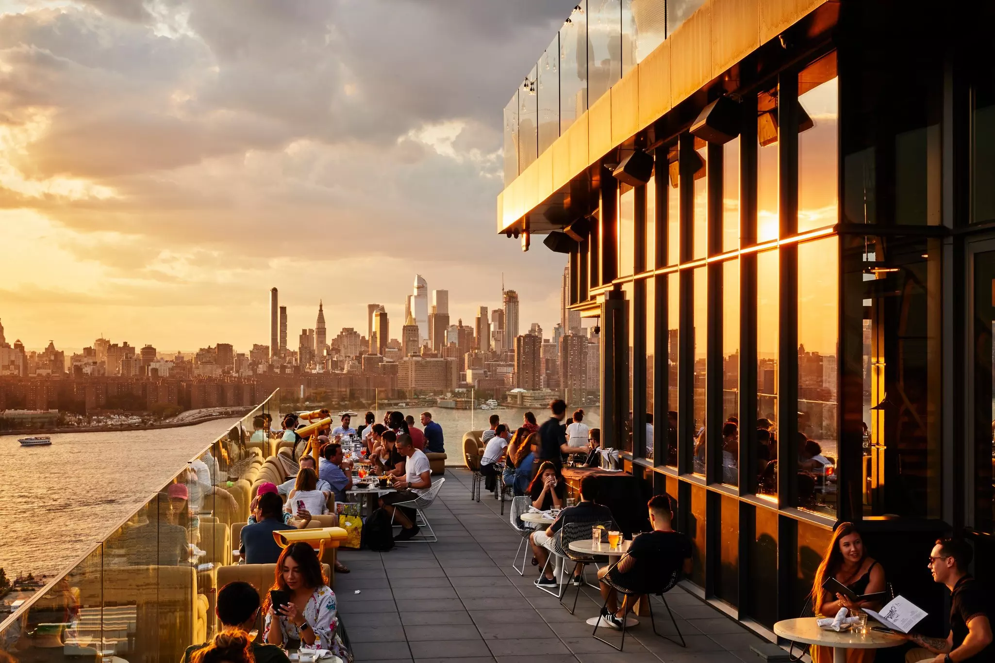People sit at tables on a roofdeck in early evening; a city skyline is across a river from the venue.