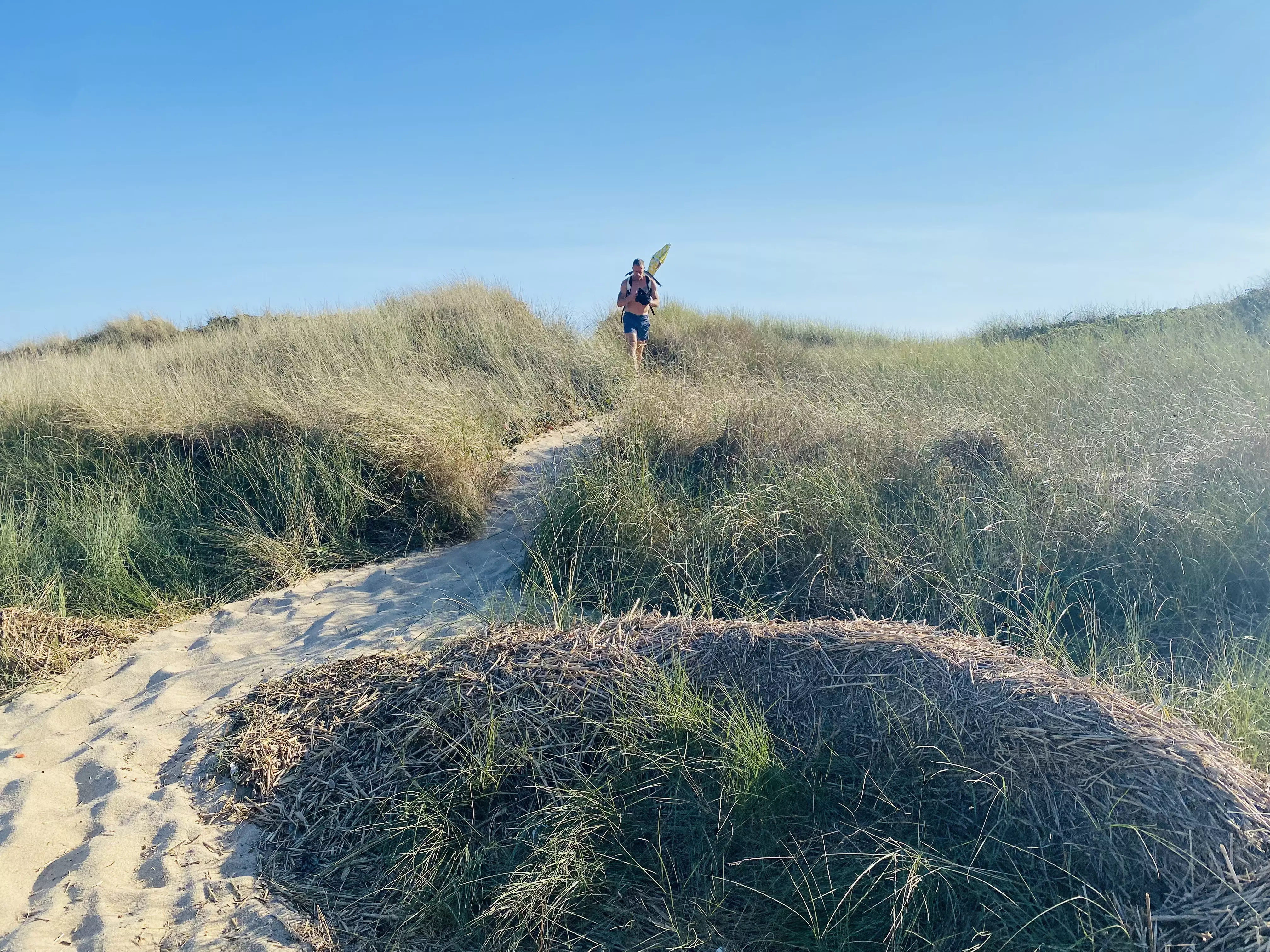 A man carrying a backpack walks along a sandy path among grass-topped dunes.