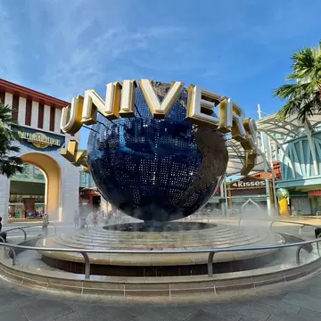 Large blue Universal globe with gold lettering above a circular fountain, flanked by palm trees and an entrance arch.