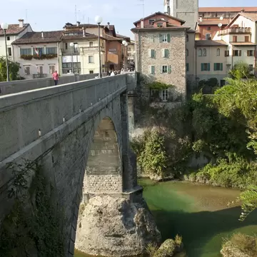 Stone arched bridge over a clear green river in Cividale del Friuli, with pastel houses and a stone bell tower above.