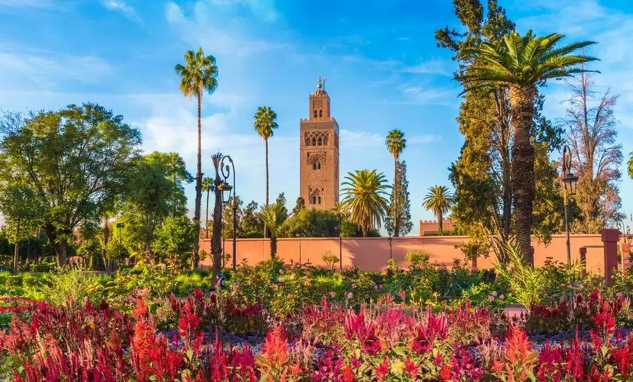 Koutoubia Mosque minaret in Marrakesh rising above palms and a pink wall, with red flowerbeds in the foreground