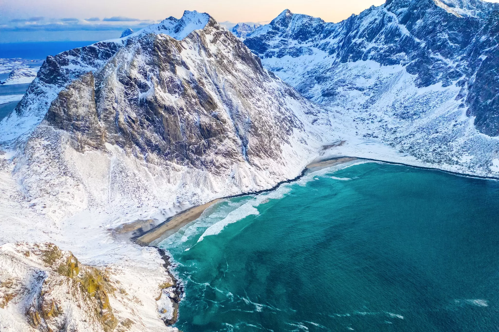 Blue-green waters lap a beach shore mountains covered in snow around it