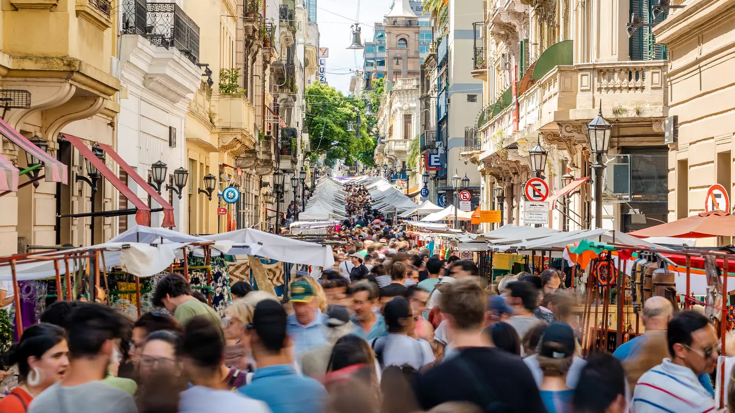 An open market takes place on a city street. The market goes on for several blocks, and the street is closed to cars and crowded with shoppers, vendors and pedestrians.