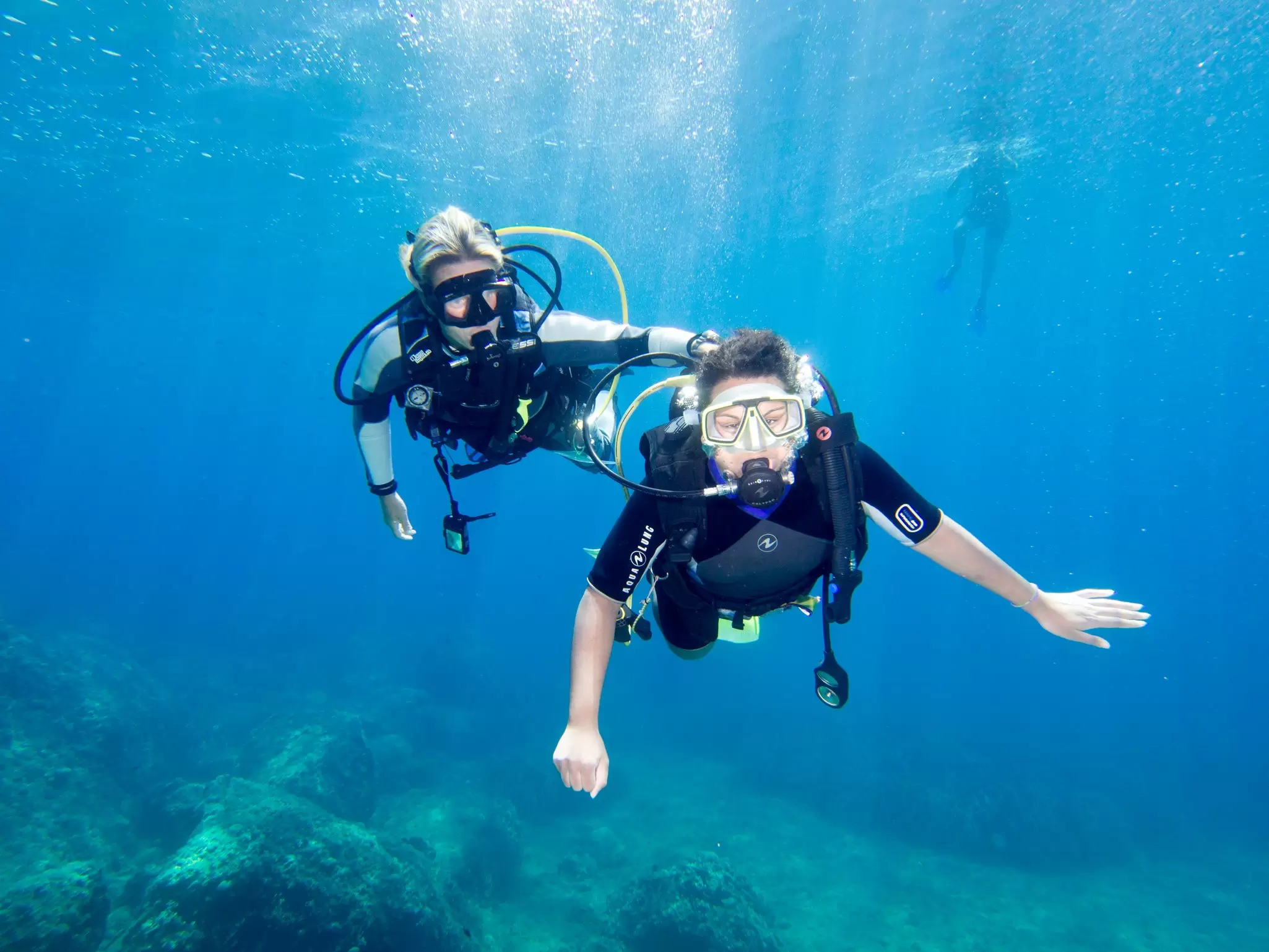 A scuba diver is supported by a diving instructor as they swim through clear water at depth in Greece