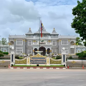 Grey Beaux-Arts palace with gilded details and a Lao flag behind an ornate gate and sign