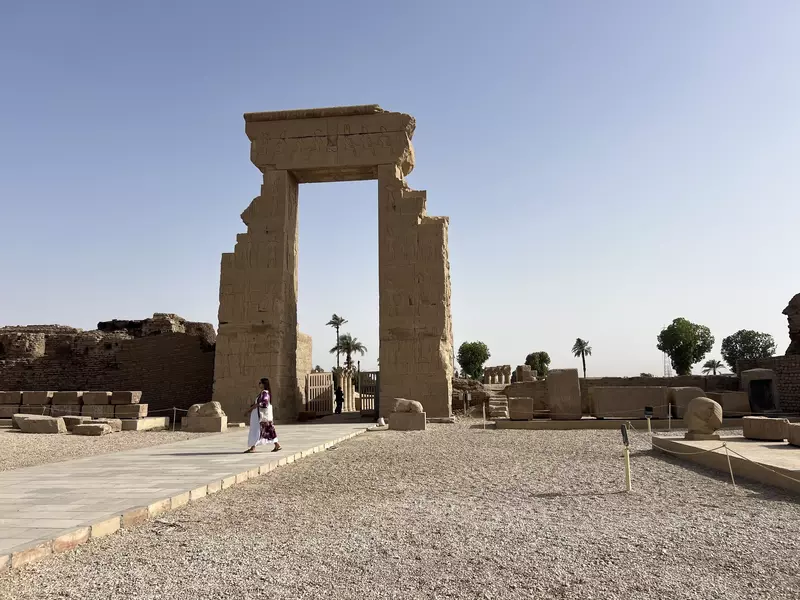 A stone archway decorated with ancient art through which you can see two palm trees. A woman walks in front