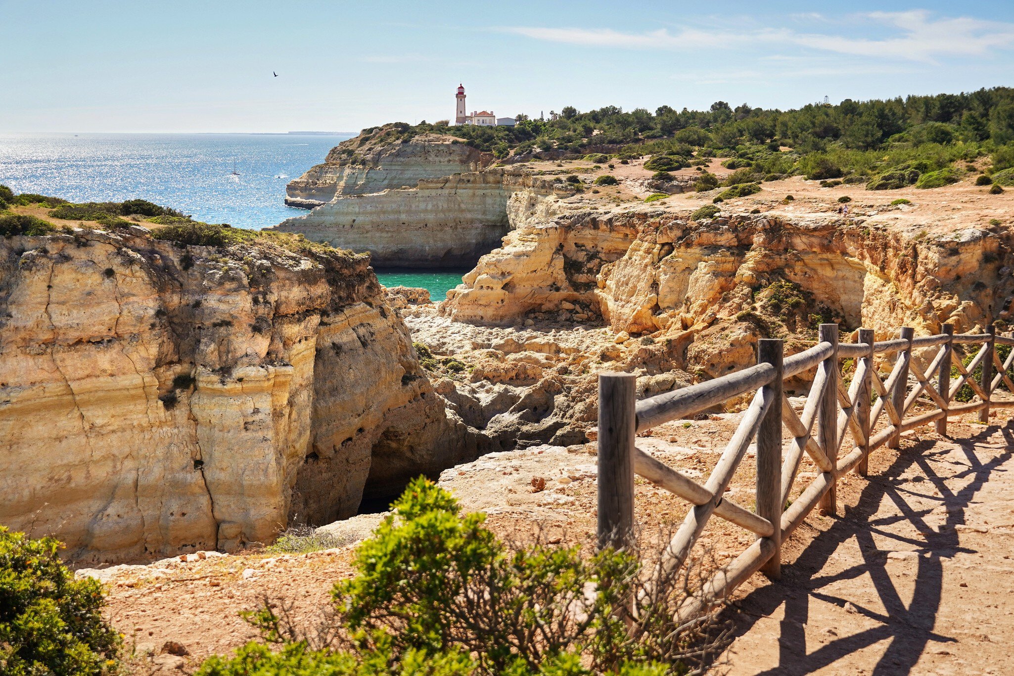 A wooden rail separates a dirt path from golden cliffs by the ocean; a white lighthouse with a red roof is in the distance.