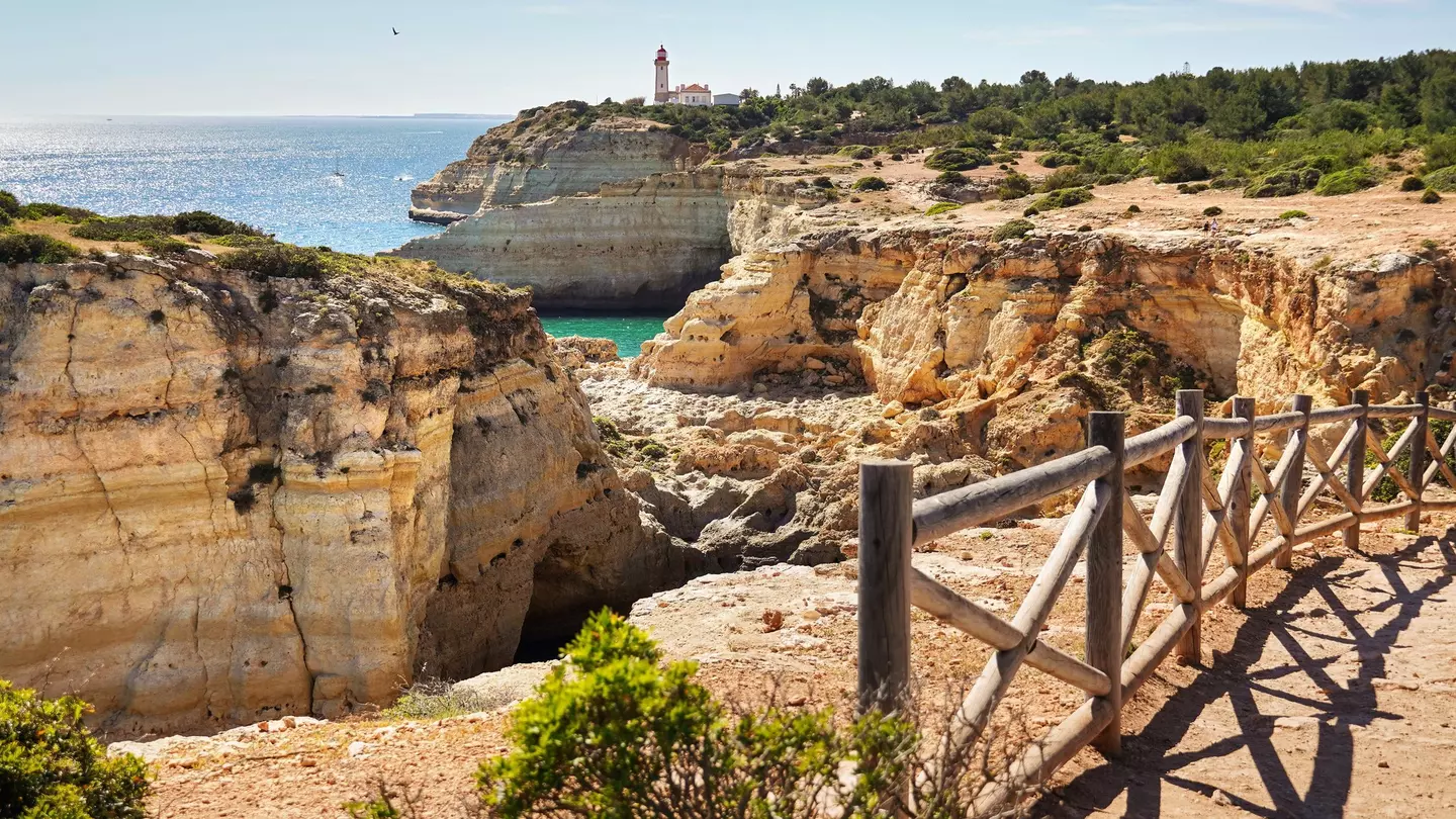 A wooden rail separates a dirt path from golden cliffs by the ocean; a white lighthouse with a red roof is in the distance.