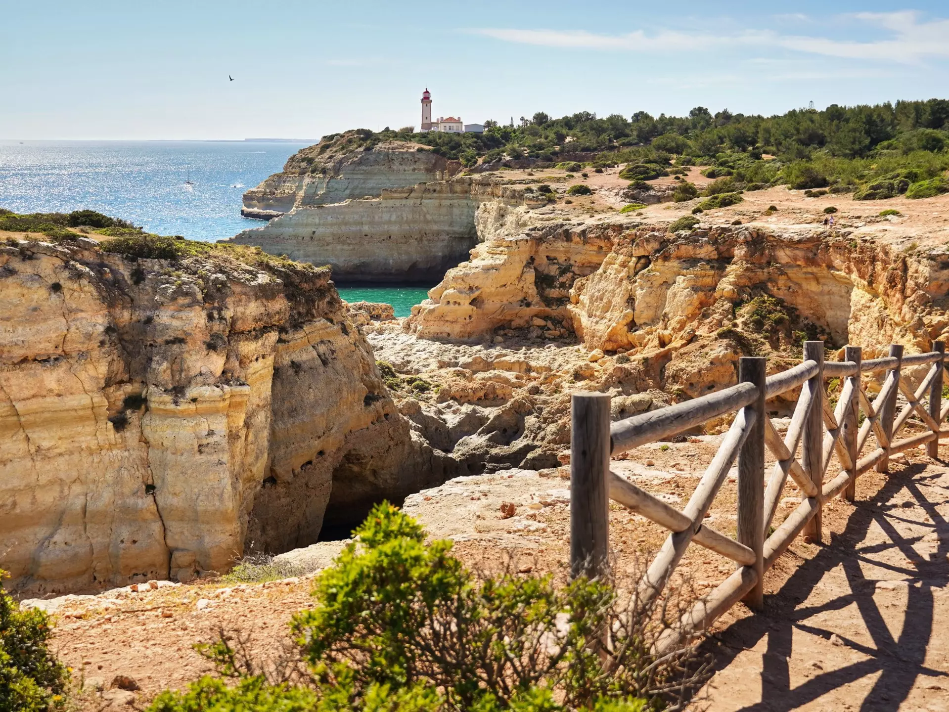 A wooden rail separates a dirt path from golden cliffs by the ocean; a white lighthouse with a red roof is in the distance.