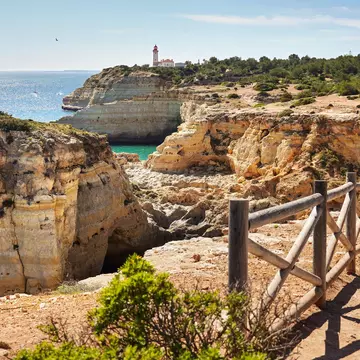 A wooden rail separates a dirt path from golden cliffs by the ocean; a white lighthouse with a red roof is in the distance.