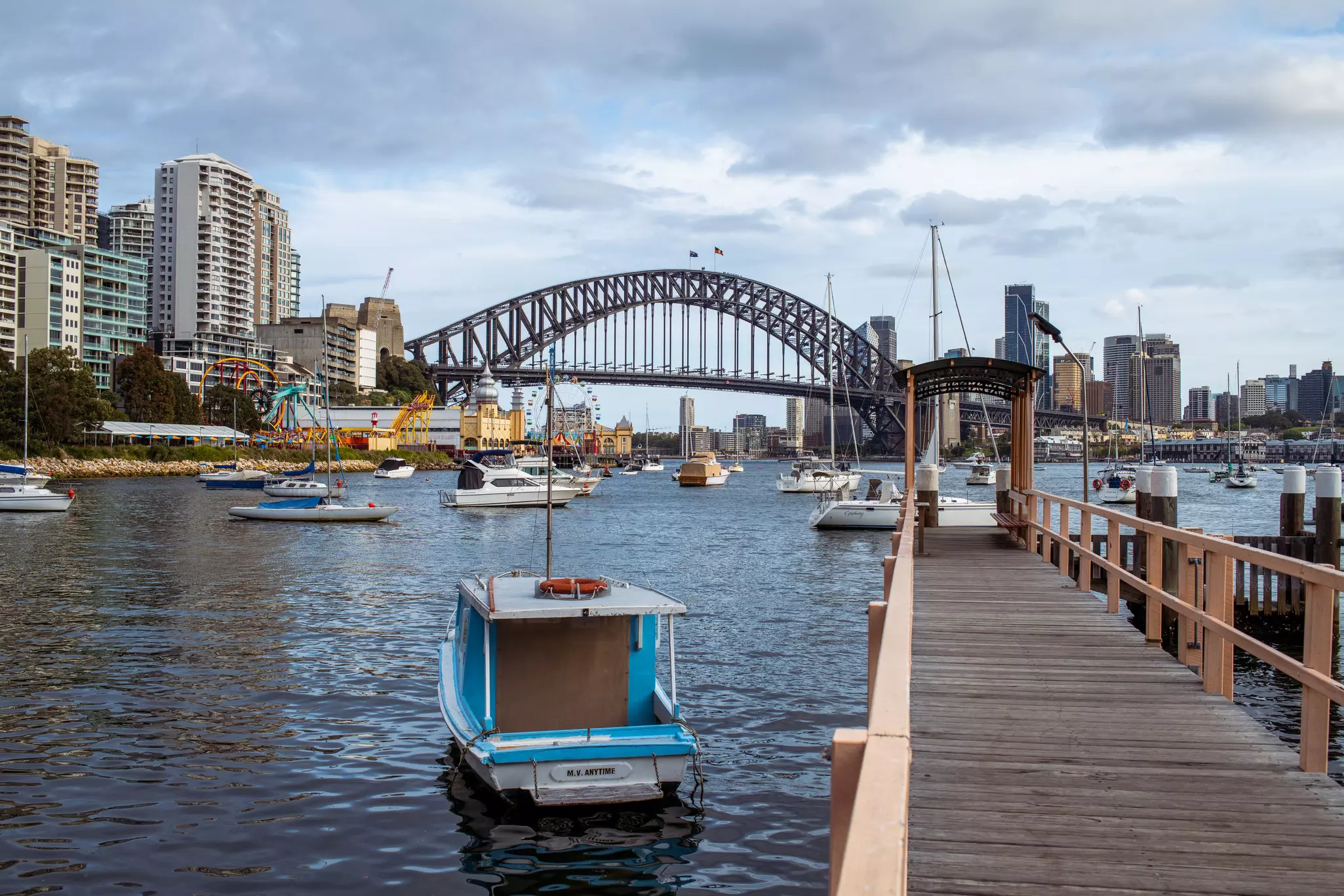 Harbor with moored boats, a wooden walkway and a city skyline with bridge crossing the water in the distance on a cloudy day.