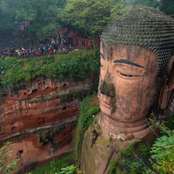 Giant stone Buddha head carved into a red sandstone cliff with ferns, crowds of people on a path above, at Leshan, China.