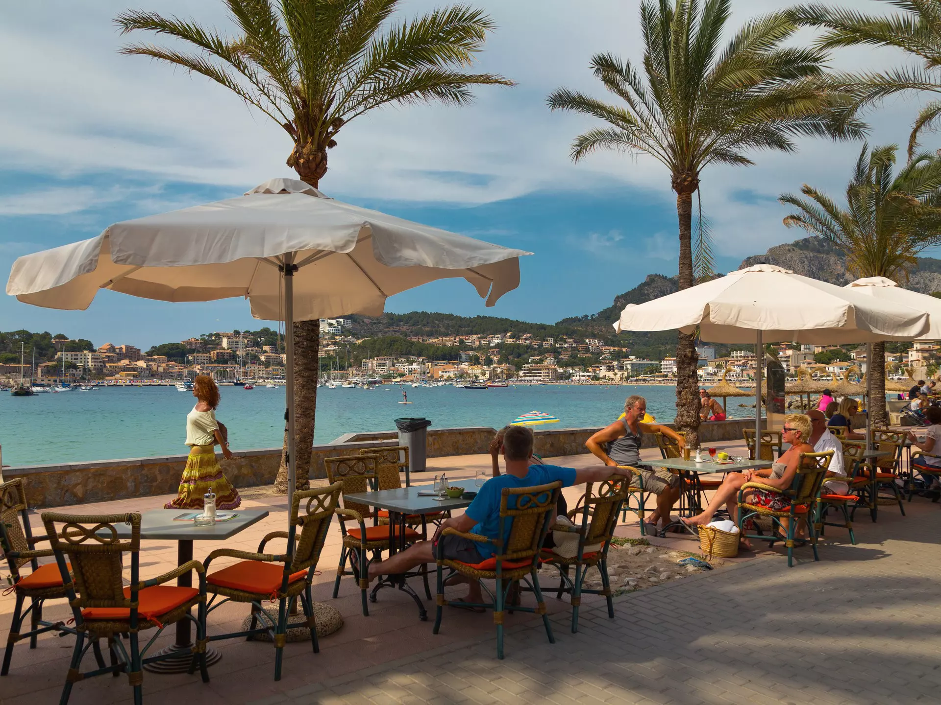 People sit at tables under palm trees by a promenade next to the sea.
