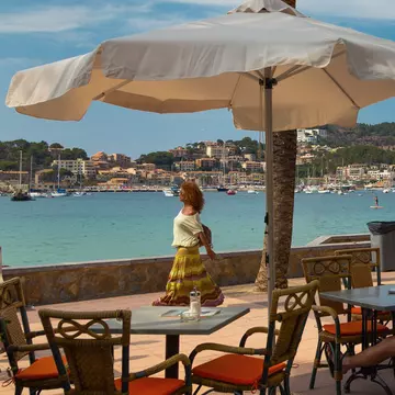 People sit at tables under palm trees by a promenade next to the sea.